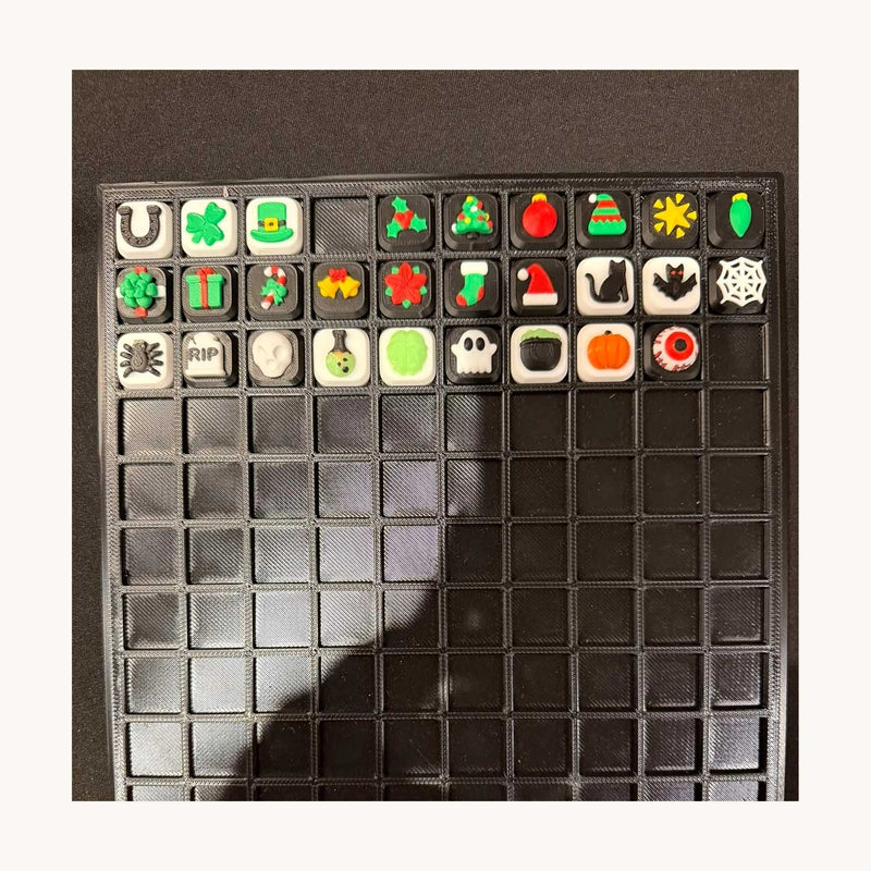 Display tray of 3D-printed square holiday and Halloween charms — shamrocks, gifts, a tree, a pumpkin, a ghost and a spider web