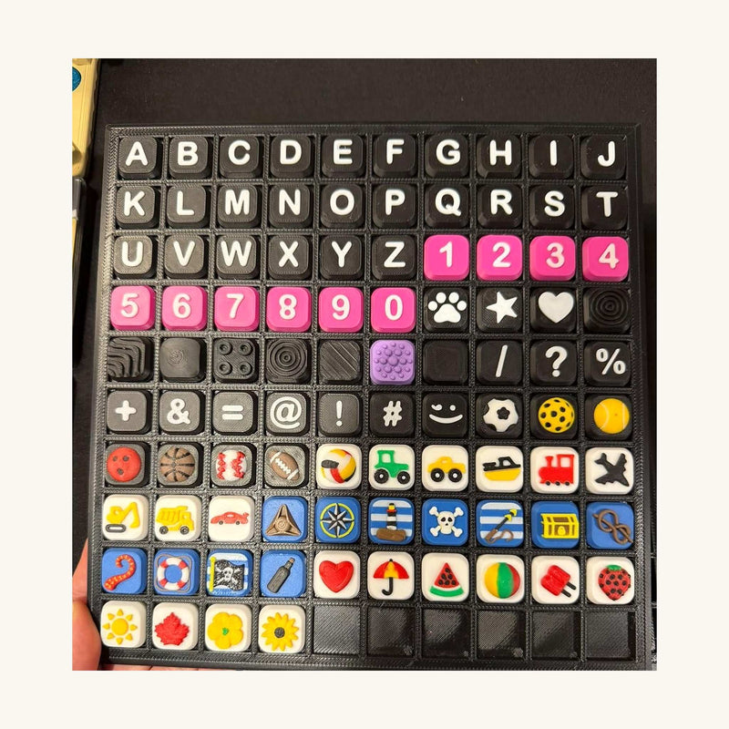 Display tray of 3D-printed square charms with raised letters, numbers, hearts, paw prints, vehicles and fruit